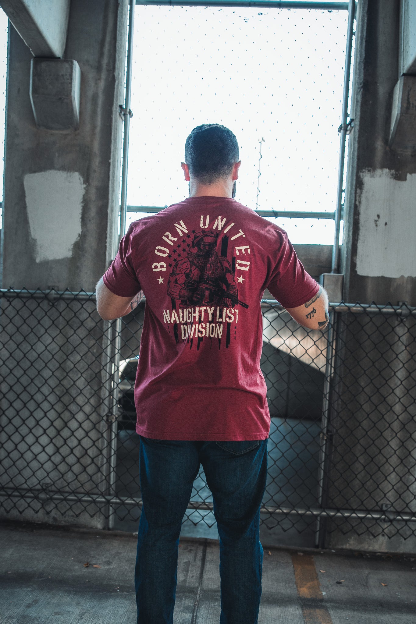 A person stands with their back to the camera, wearing a Born United Private Label Tactical Santa Tee and blue jeans, in front of a chain-link fence and concrete pillars.