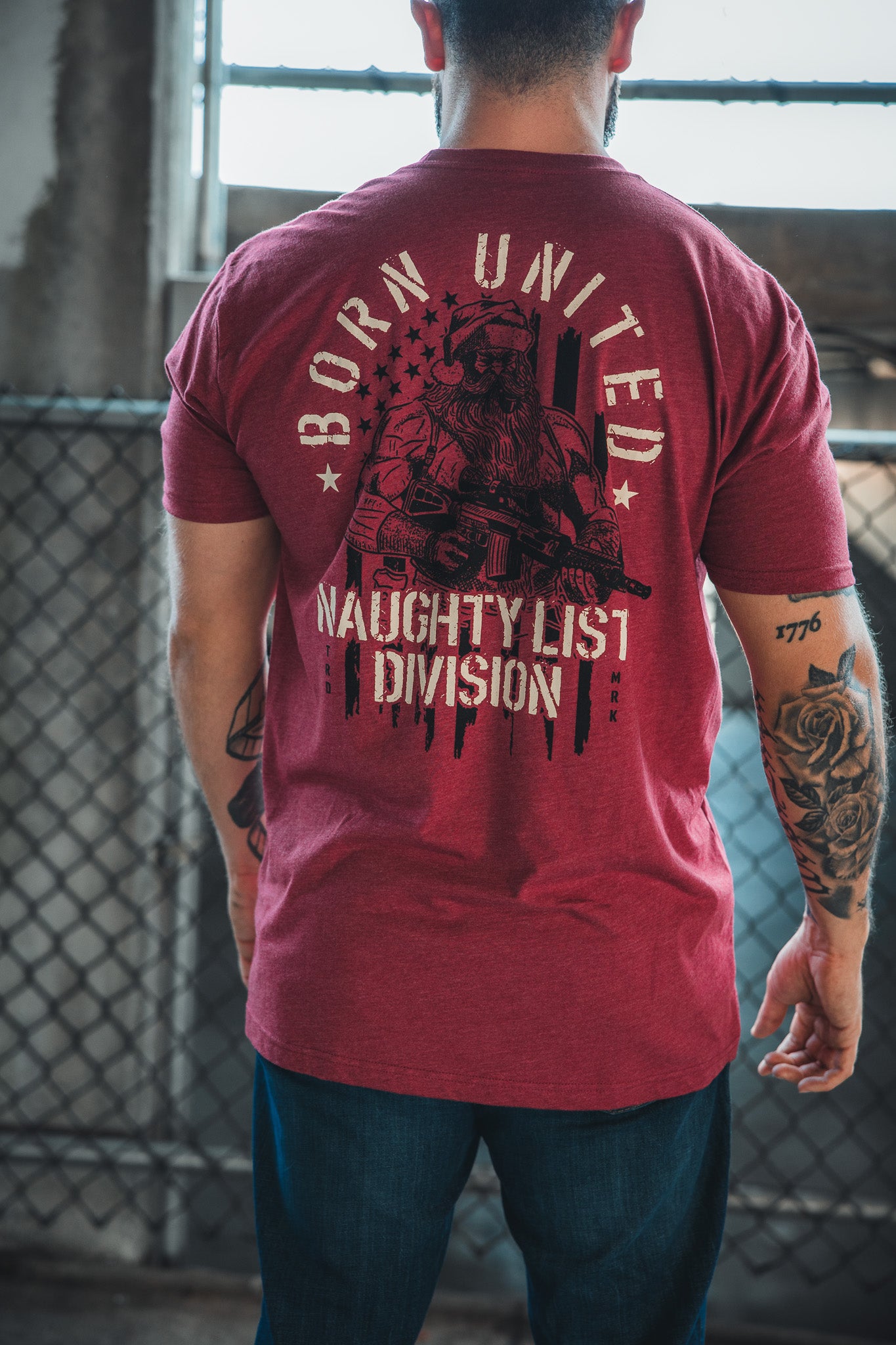 A person stands with their back to the camera, sporting the Born United Private Label Tactical Santa Tee, which features an armed Saint Nick in sunglasses over an American flag and reads "Born United Naughty List Division"—ideal for Christmas cheer.