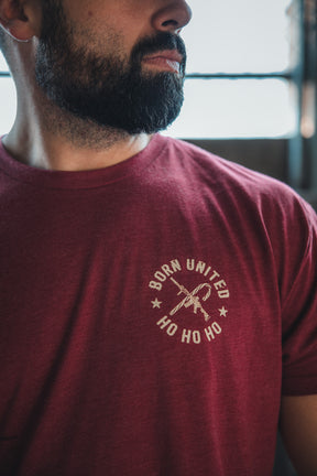 A bearded man wears the Born United Private Label Tactical Santa Tee, a maroon limited-edition shirt featuring a rifle graphic and "BORN UNITED HO HO HO." The image is cropped to show his partial face and upper torso.