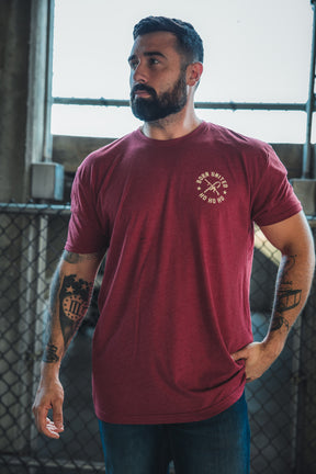 A bearded man stands indoors near a chain-link fence, wearing the Born United Private Label Tactical Santa Tee—featuring "Ho Ho Ho" and crossed axes—paired with blue jeans and displaying arm tattoos as he looks to the side.
