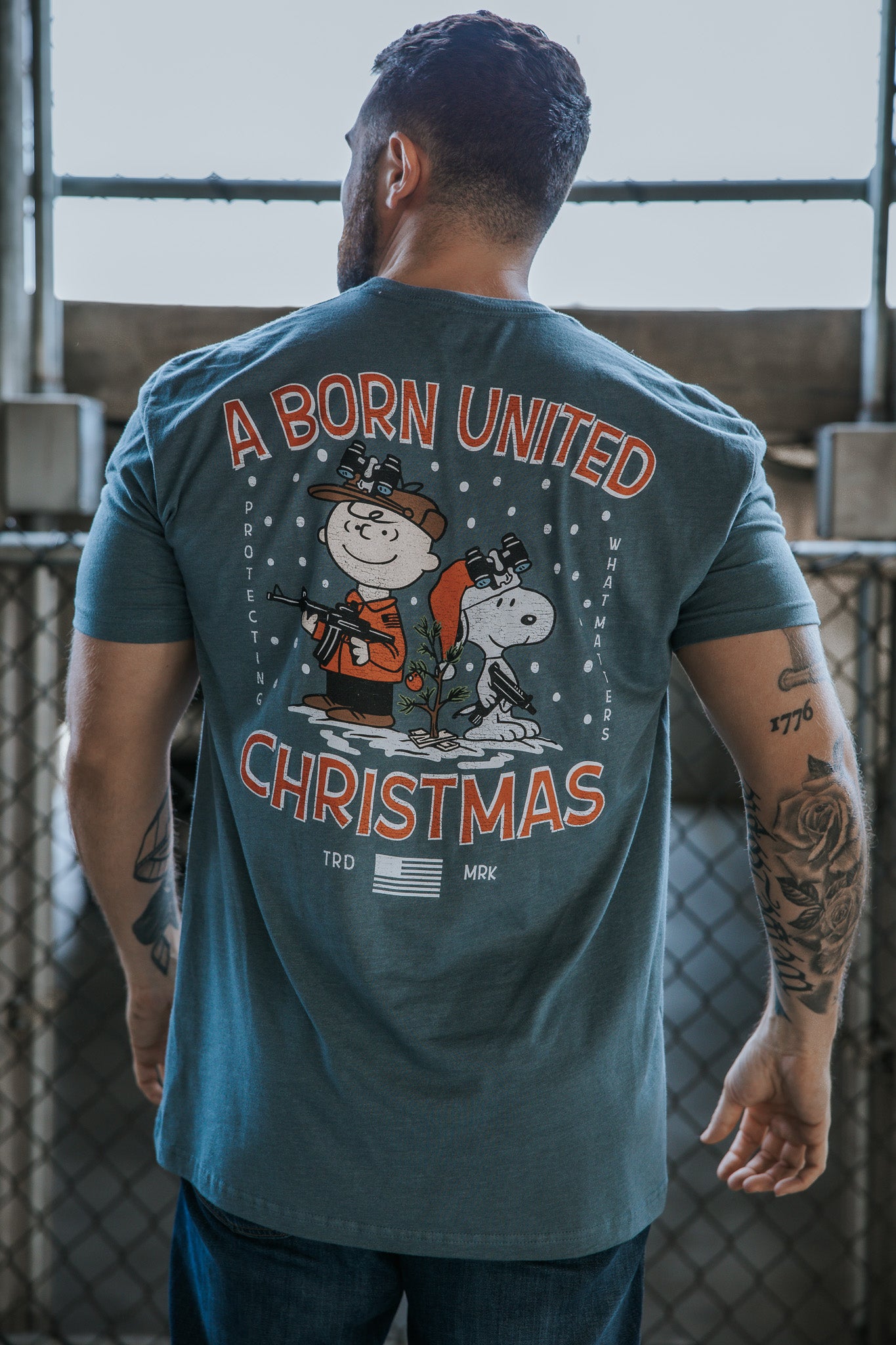 A man faces away from the camera in a Born United Private Label tee, “A Gnarly Brown Christmas,” featuring Snoopy, Charlie Brown in festive attire, additional text, and a U.S. flag—perfect for the holiday season.