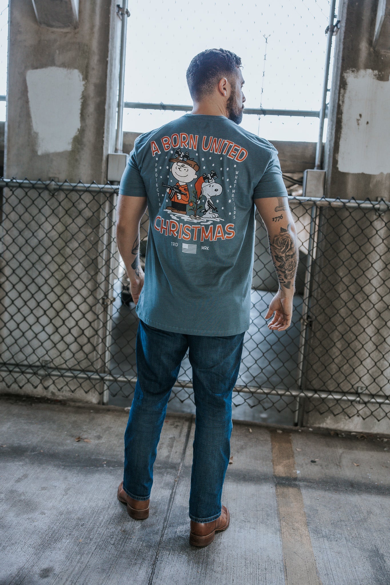 A man stands outdoors with his back to the camera, wearing jeans and a blue retro-inspired Born United Private Label tee featuring a Peanuts Christmas graphic and the text "A Gnarly Brown Christmas.