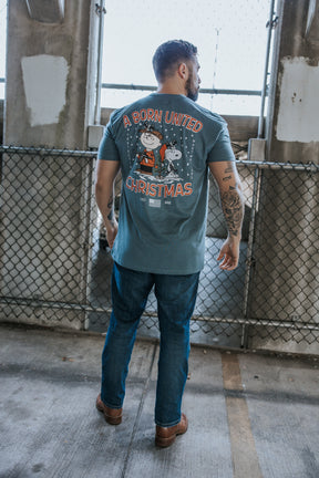 A man stands outdoors with his back to the camera, wearing jeans and a blue retro-inspired Born United Private Label tee featuring a Peanuts Christmas graphic and the text "A Gnarly Brown Christmas.