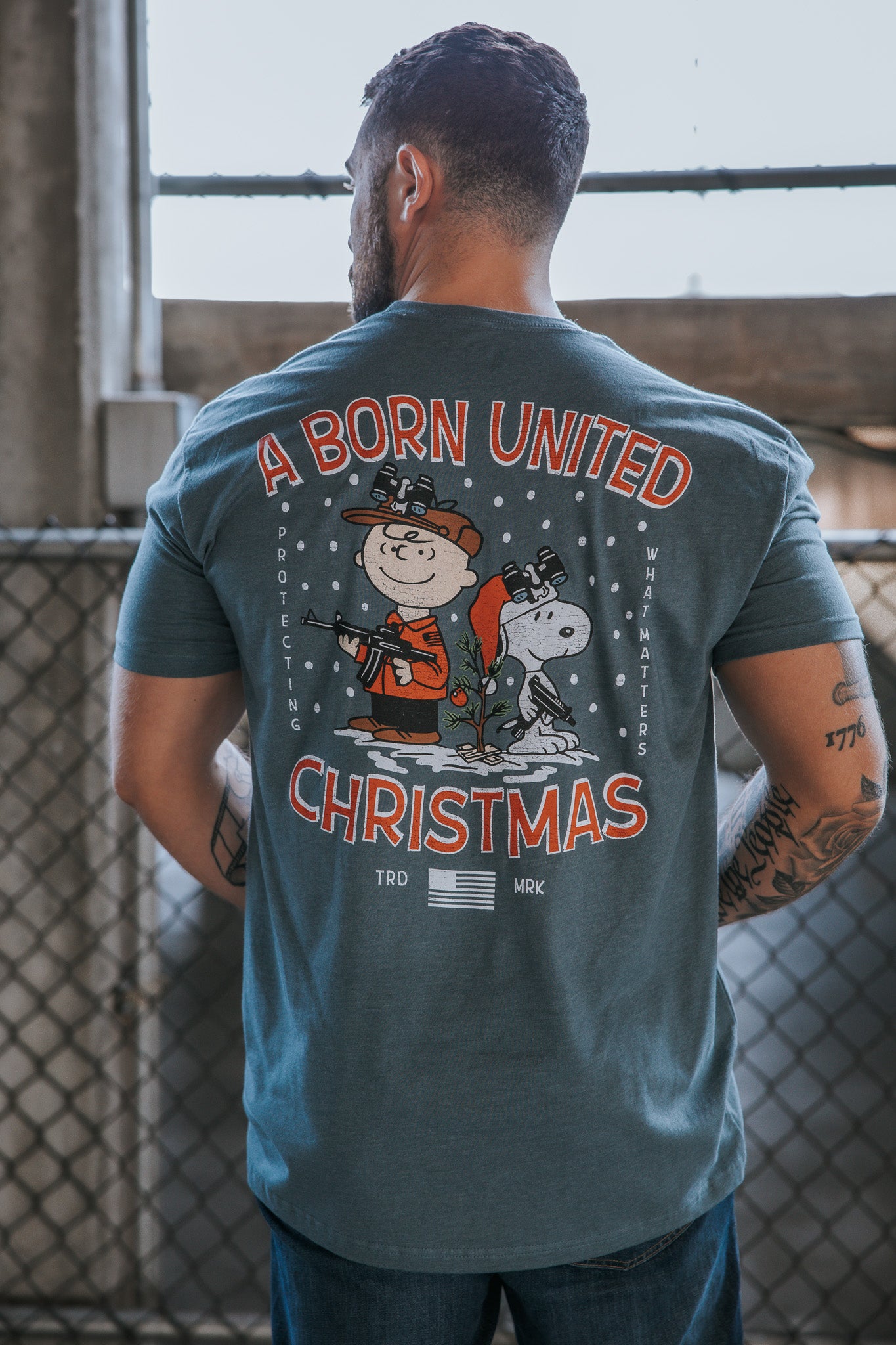 A man stands with his back to the camera, wearing Born United Private Label’s “A Gnarly Brown Christmas” tee, featuring Charlie Brown and Snoopy with festive decorations for classic Peanutty holiday vibes.
