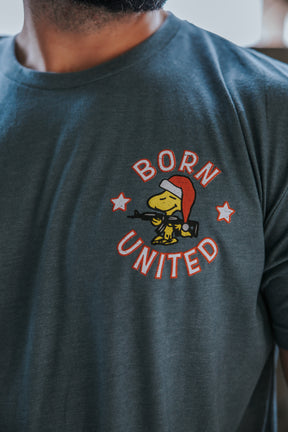 A person wears the "A Gnarly Brown Christmas" tee by Born United Private Label, featuring a yellow cartoon bird in a Santa hat holding a rifle with "BORN UNITED" and two stars—a playful, Peanut-inspired holiday design.
