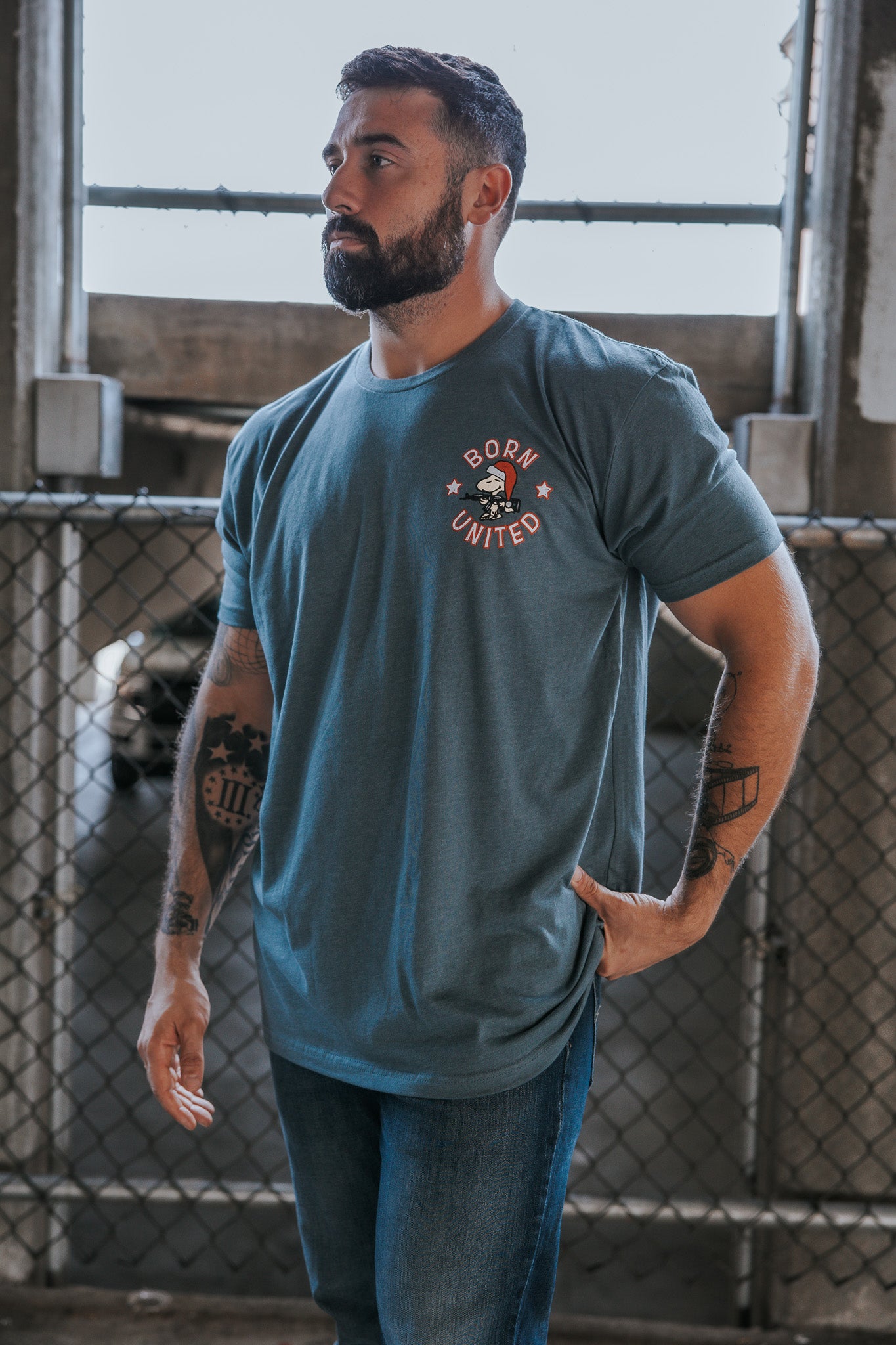A bearded, tattooed man stands by a chain-link fence, wearing jeans and the Born United Private Label "A Gnarly Brown Christmas" tee. He looks to the side with one hand on his hip.