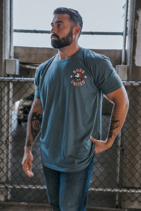 A bearded, tattooed man stands by a chain-link fence, wearing jeans and the Born United Private Label "A Gnarly Brown Christmas" tee. He looks to the side with one hand on his hip.