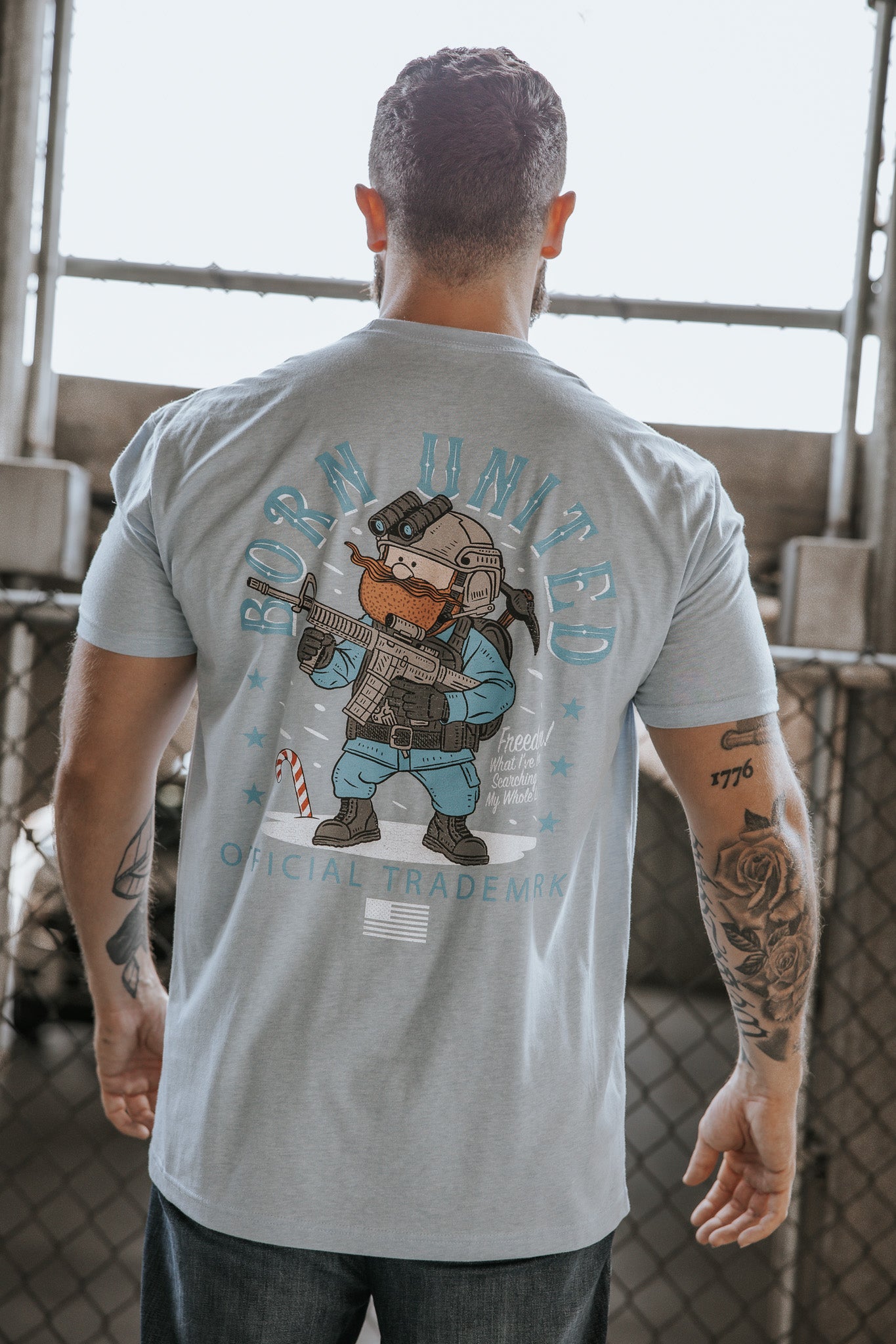 A tattooed man wears the Yukon Cornelius Tee by Born United Private Label, a light blue shirt with a cartoon bear in tactical gear and "Born United Official Trademark" on the back, standing before a chain-link fence—celebrating individuality.
