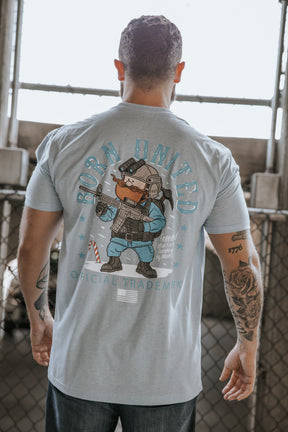A tattooed man wears the Yukon Cornelius Tee by Born United Private Label, a light blue shirt with a cartoon bear in tactical gear and "Born United Official Trademark" on the back, standing before a chain-link fence—celebrating individuality.