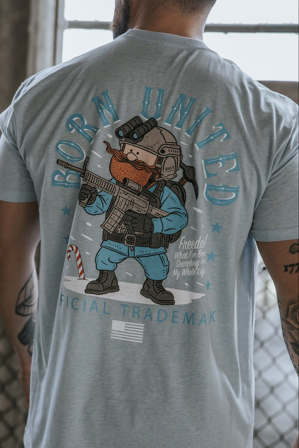 A tattooed person wears the Yukon Cornelius Tee from Born United Private Label—a light blue shirt with a cartoon bearded soldier holding a rifle, stars, and “Born United,” “Official Trademark,” symbolizing freedom.