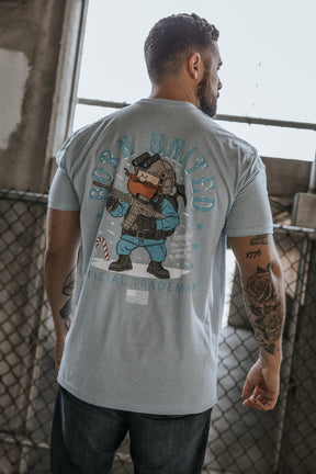 A man stands with his back to the camera, wearing the Yukon Cornelius Tee from Born United Private Label, which features a cartoon soldier, stars, and "BORN UNITED" on the back, beside a chain-link fence in an industrial setting.