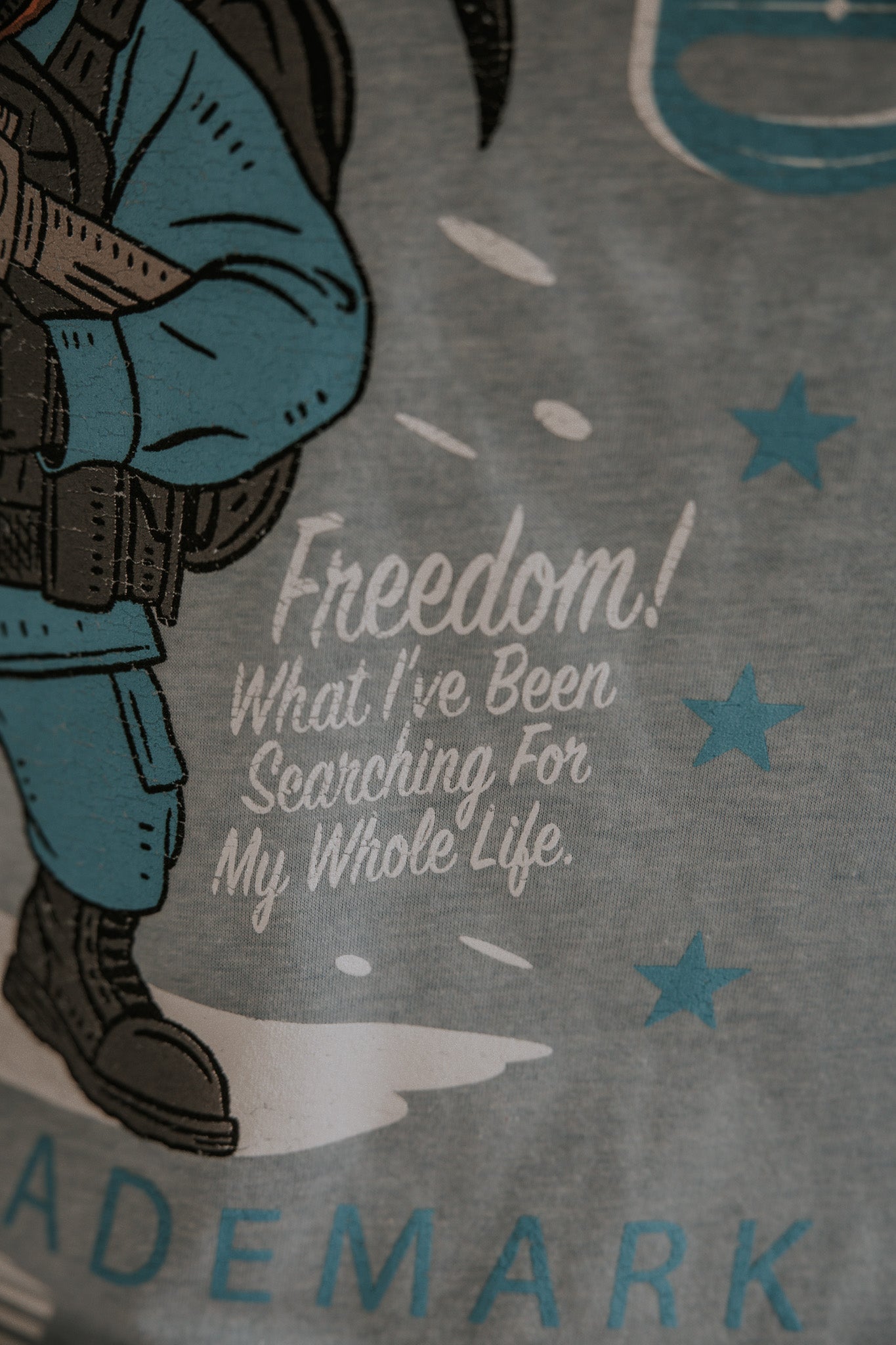 A close-up of the Yukon Cornelius Tee by Born United Private Label, featuring a person in blue tactical gear and the text "Freedom! What I've Been Searching For My Whole Life," with blue stars highlighting the freedom theme.