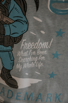 A close-up of the Yukon Cornelius Tee by Born United Private Label, featuring a person in blue tactical gear and the text "Freedom! What I've Been Searching For My Whole Life," with blue stars highlighting the freedom theme.