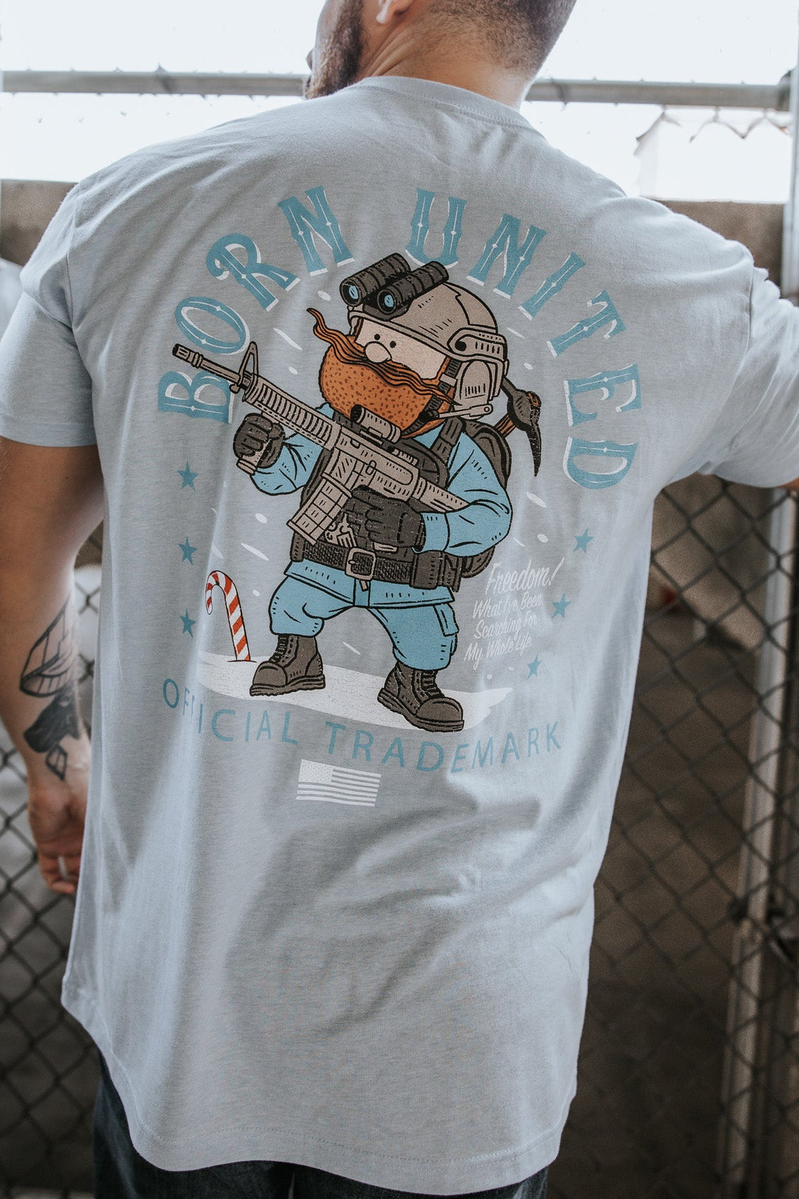 A man stands with his back turned, wearing the Yukon Cornelius Tee by Born United Private Label. The light blue shirt features a bear in tactical gear and the text "BORN UNITED" and "OFFICIAL TRADEMARK." A chain-link fence is behind him.
