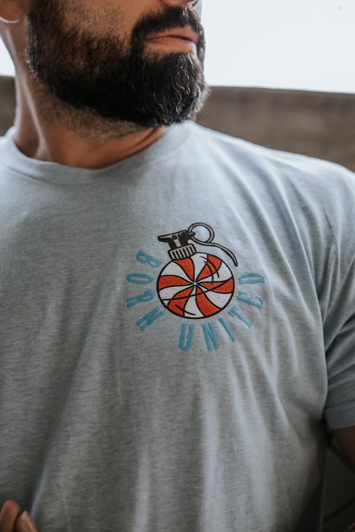A bearded man wears the Yukon Cornelius Tee by Born United Private Label—a light blue shirt with a red and white striped grenade graphic and "BORN UNITED" in blue, representing unity and freedom.
