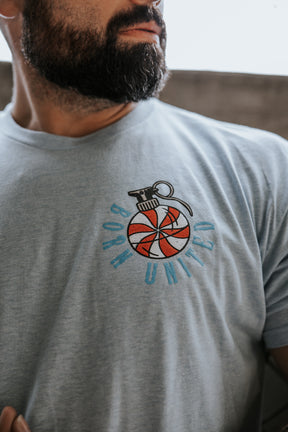 A bearded man wears the Yukon Cornelius Tee by Born United Private Label—a light blue shirt with a red and white striped grenade graphic and "BORN UNITED" in blue, representing unity and freedom.