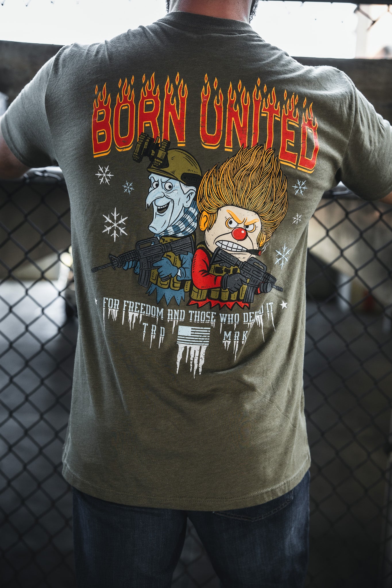 Someone wears a green Born United Private Label "Tactical Miser Operations" T-shirt, featuring BORN UNITED in flaming letters, cartoon characters with guns, snowflakes and icicles, and the text "FOR FREEDOM AND THOSE WHO DEAL IT" below.