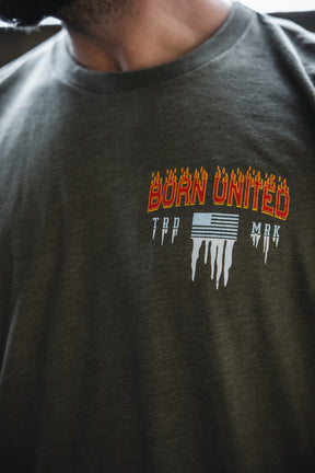 A person wearing a green Tactical Miser Operations t-shirt by Born United Private Label, featuring "BORN UNITED" in red with flames above a melting American flag and TRD MRK lettering.