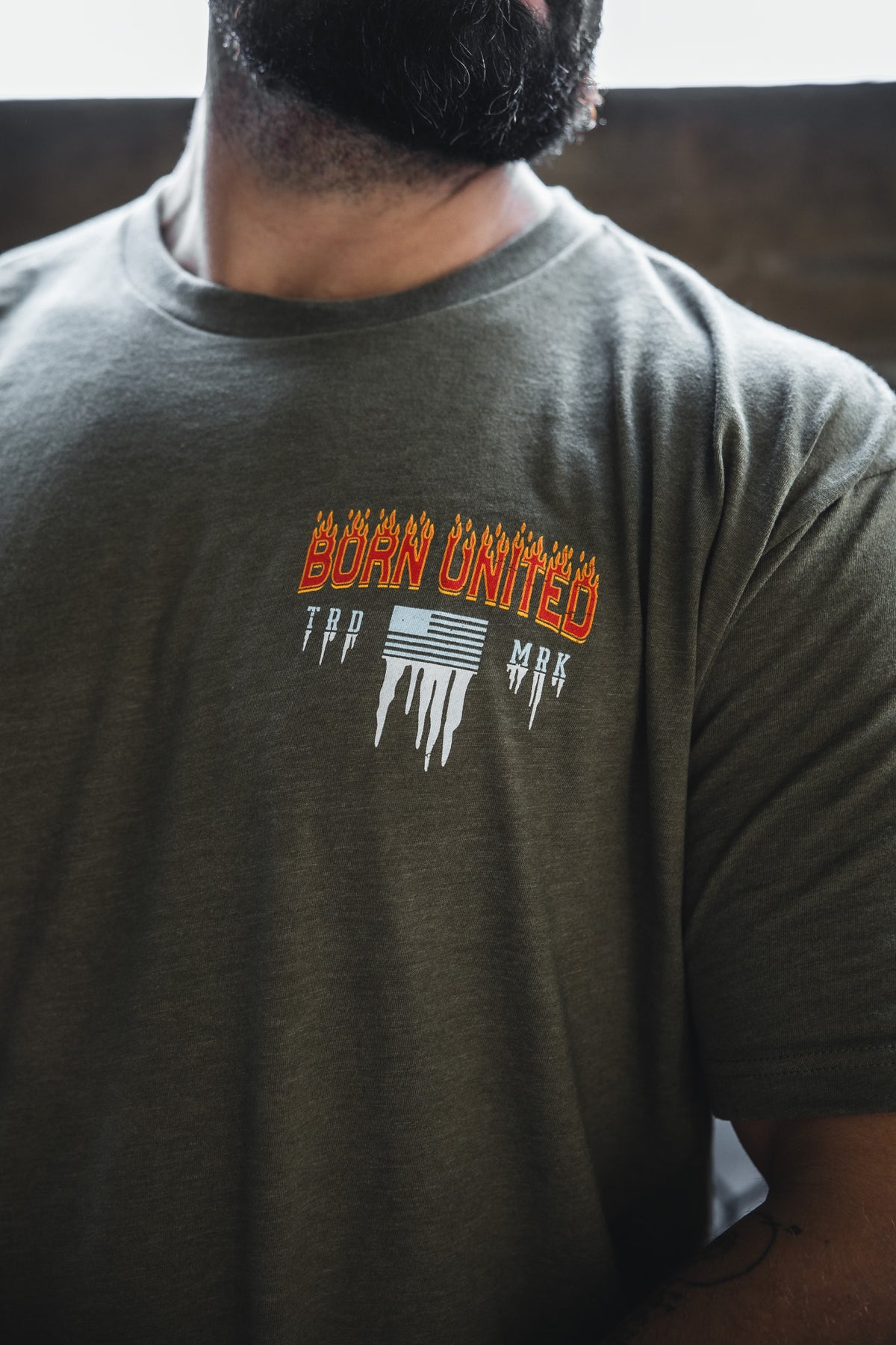 A man wears an olive green Tactical Miser Operations t-shirt by Born United Private Label, featuring “BORN UNITED” in orange flaming letters with white melting lines and text on the left chest.