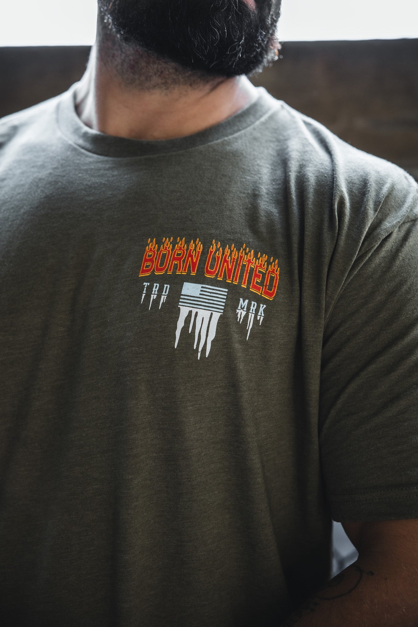A man wears an olive green Tactical Miser Operations t-shirt by Born United Private Label, featuring “BORN UNITED” in orange flaming letters with white melting lines and text on the left chest.