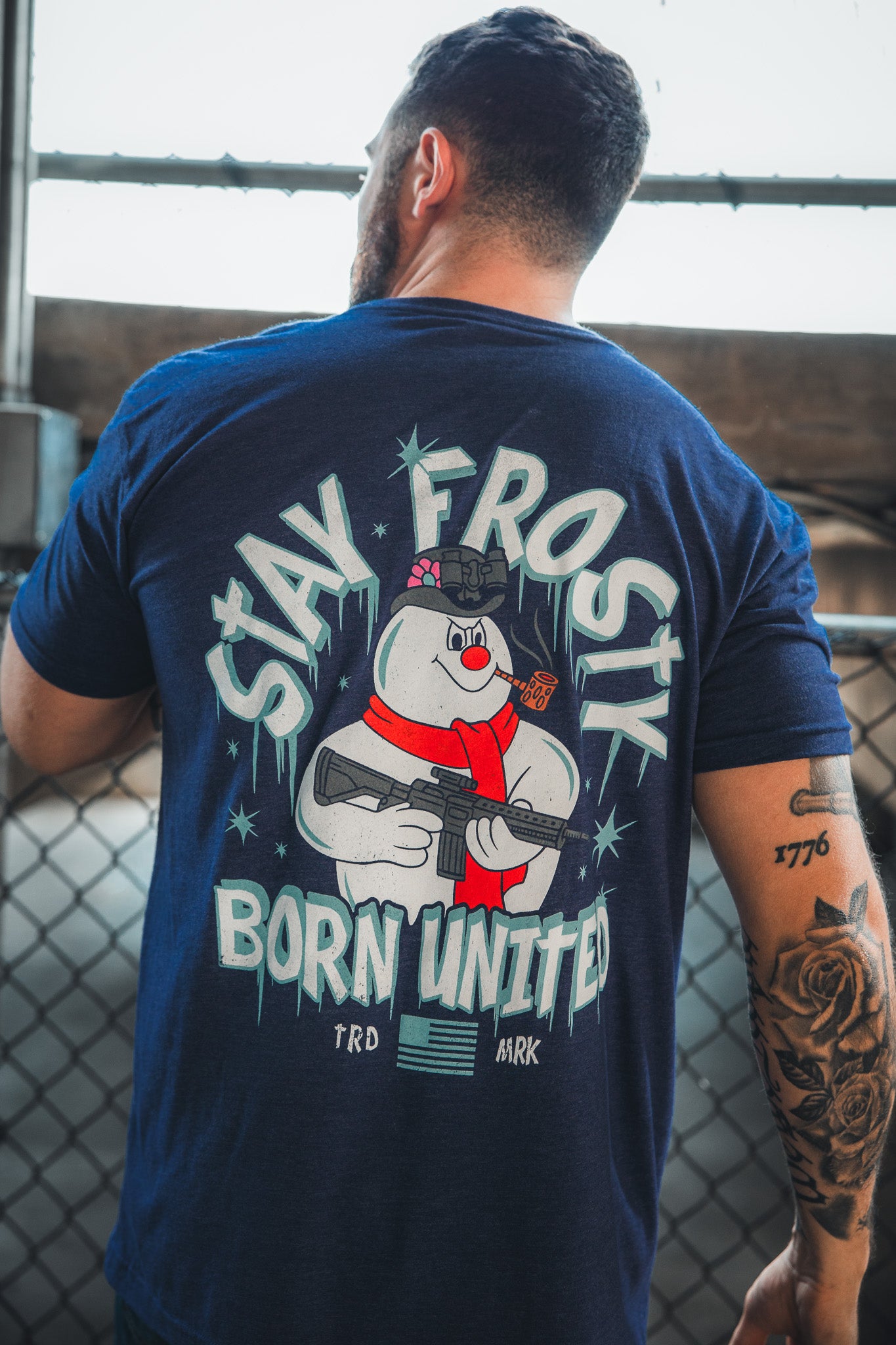 A tattooed man stands by a chain-link fence, sporting the Born United Private Label “Stay Frosty” tee with a cartoon snowman in a helmet holding a rifle and patriotic “STAY FROSTY BORN UNITED” text on the back.