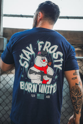 A tattooed man stands by a chain-link fence, sporting the Born United Private Label “Stay Frosty” tee with a cartoon snowman in a helmet holding a rifle and patriotic “STAY FROSTY BORN UNITED” text on the back.