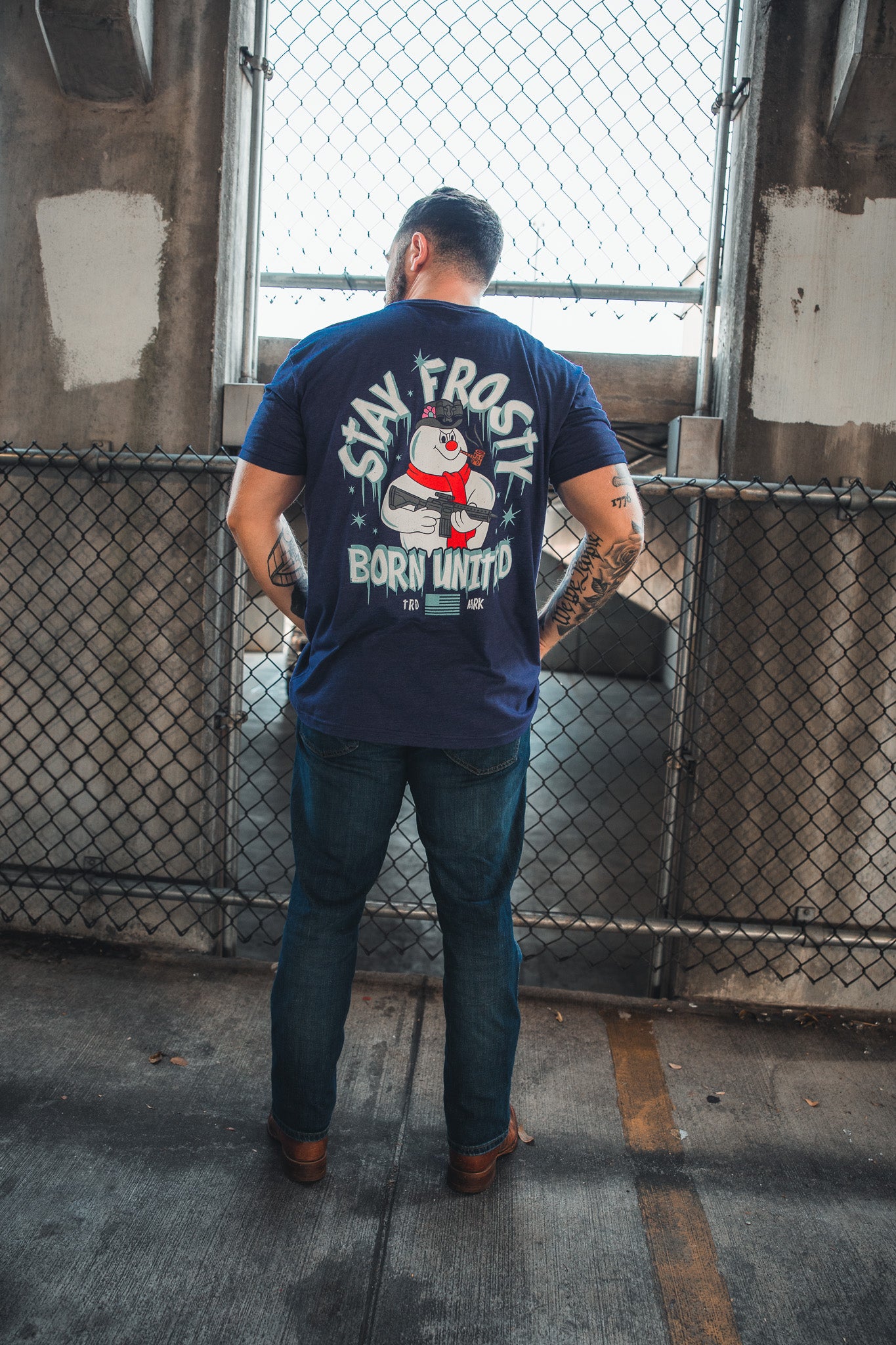 A man faces away from the camera in front of a chain-link fence, wearing Born United Private Label’s navy blue Stay Frosty tee featuring “STAY FROSTY BORN UNITED” and a patriotic Frosty graphic making a hand gesture.
