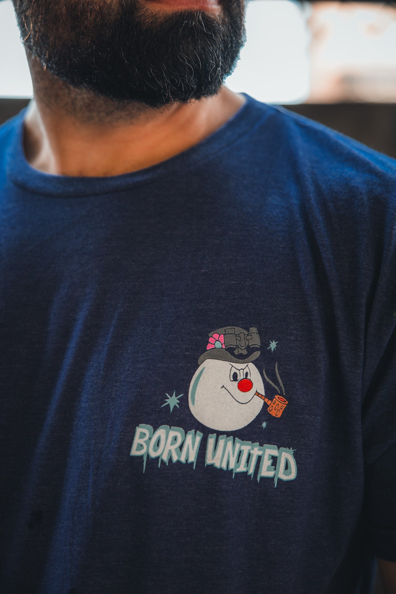 A bearded man wears the Stay Frosty tee by Born United Private Label: a navy retro shirt with a cartoon snowman in a pink bow and smoking pipe, featuring the text “BORN UNITED” below—perfect holiday style.