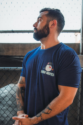 A bearded, tattooed man stands by a chain-link fence, wearing a retro "Stay Frosty" tee from Born United Private Label with a patriotic Frosty snowman and BORN UNITED text on the front.