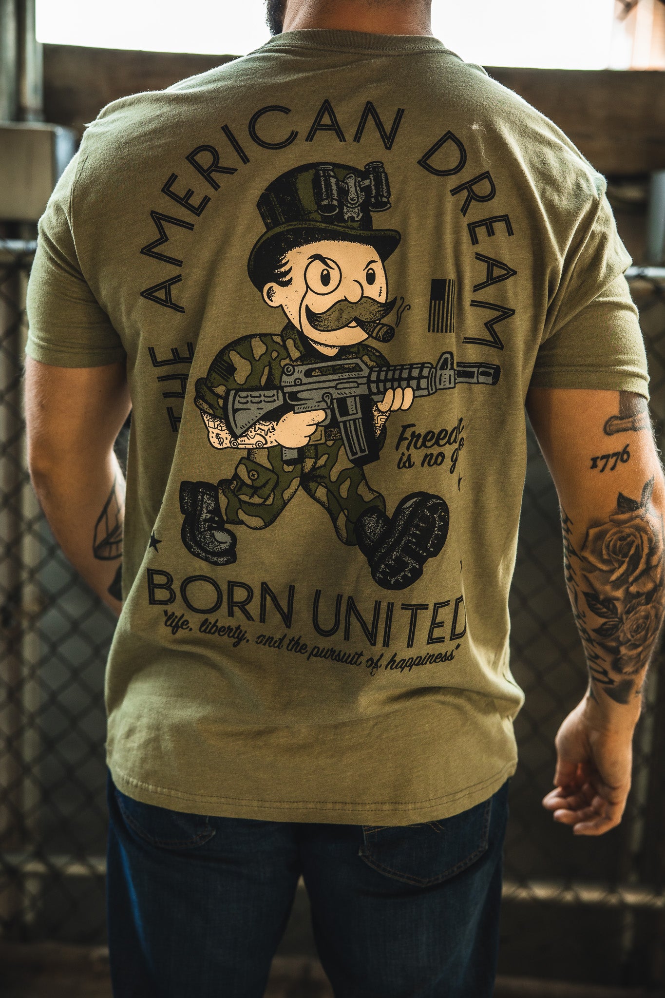 A man wears "The American Dream" olive green tee by Born United Private Label, featuring a cartoon soldier and slogan—a reminder that life, liberty, and happiness come with a price.