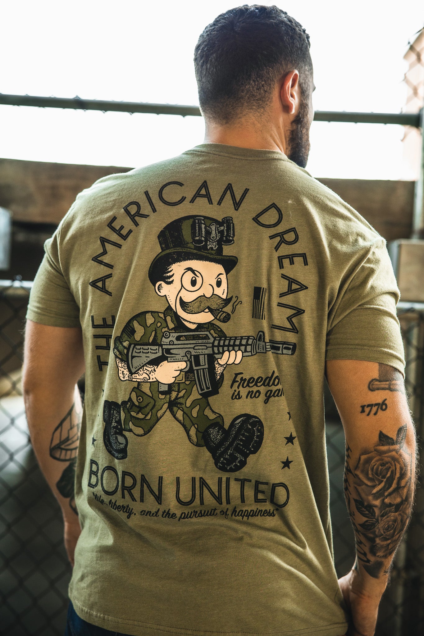 A man with tattoos wears "The American Dream" graphic tee by Born United Private Label, featuring a cartoon soldier and "freedom is no game." He stands by a chain-link fence, adding edge to his bold statement look.