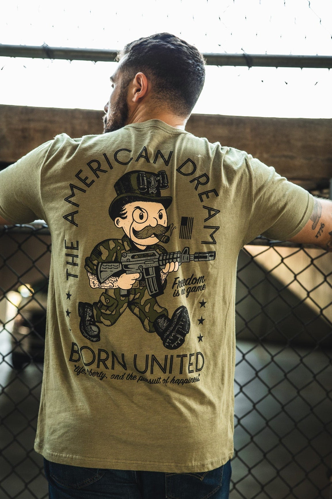 A person faces away from the camera, wearing Born United Private Label’s “The American Dream” tee with a Monopoly man cartoon soldier and rifle under “THE AMERICAN DREAM,” plus text: “BORN UNITED” and “freedom is no game.”.