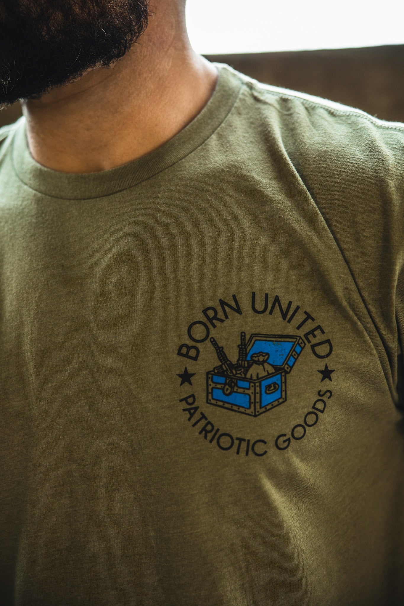 A close-up of someone in “The American Dream” t-shirt by Born United Private Label, featuring a blue chest, rifles, stars, and bold BORN UNITED PATRIOTIC GOODS branding—a strong symbol that the American Dream endures.
