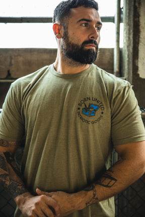 A bearded, tattooed man stands indoors wearing Born United Private Label’s "The American Dream" T-shirt—olive green with a blue "freedom is no game" graphic—looking to the side with his hands together.