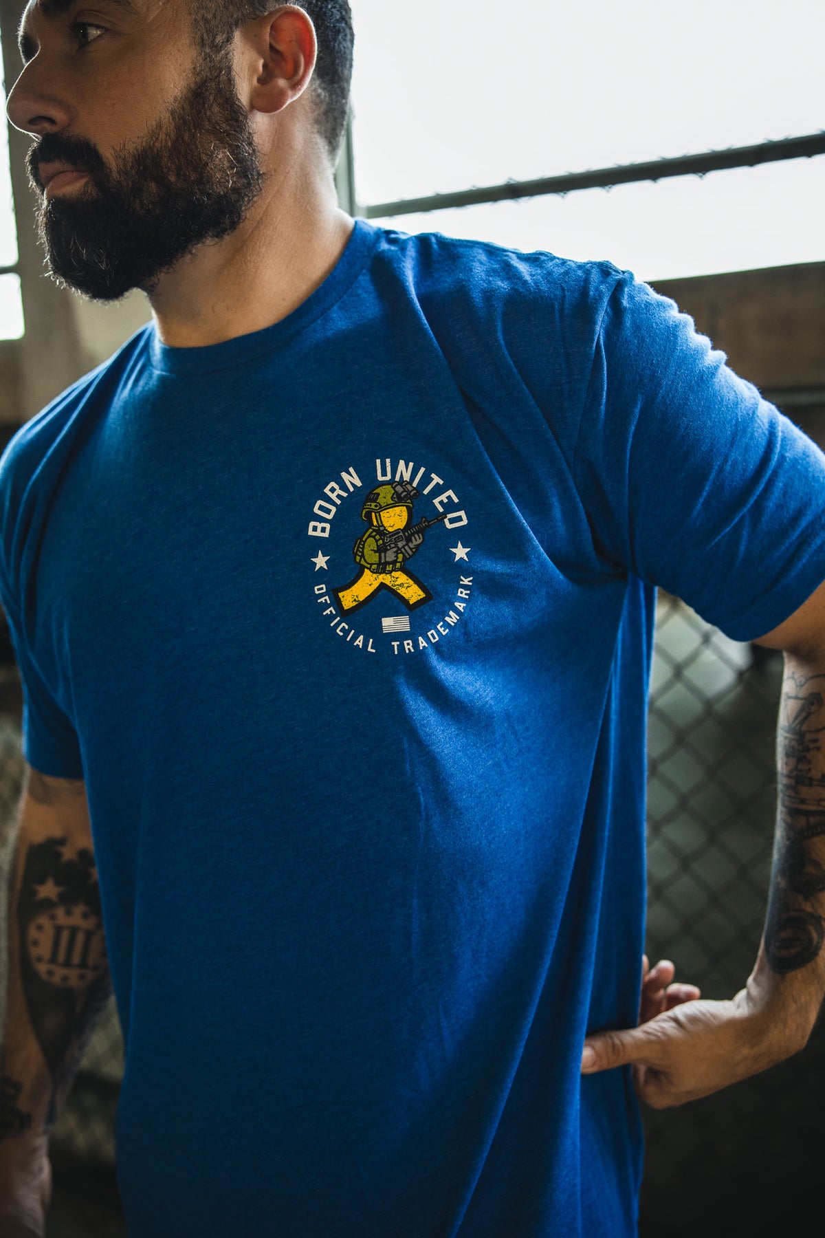 A bearded, tattooed man wears a blue "Born United Private Label" t-shirt with the "You've Got Ammo!" design, featuring a yellow cartoon character and retro tech humor, as he stands partially turned near a wire fence.