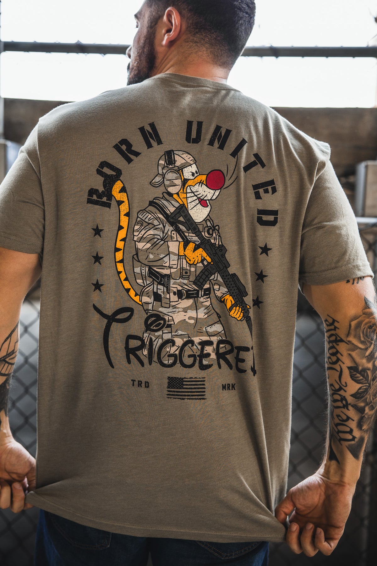 A man displays the back of his Born United Private Label "Triggered" T-shirt, featuring a cartoon soldier with a clown nose, tiger tail, rifle, stars, and the slogan "BORN UNITED TRIGGERED!" above an American flag.
