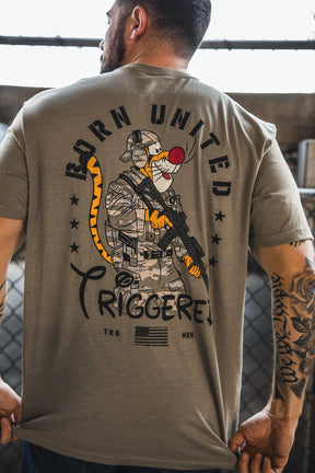 A man displays the back of his Born United Private Label "Triggered" T-shirt, featuring a cartoon soldier with a clown nose, tiger tail, rifle, stars, and the slogan "BORN UNITED TRIGGERED!" above an American flag.