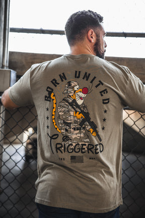 A man faces away from the camera, wearing the Born United Private Label "Triggered" khaki t-shirt with a graphic of an armed patriot sporting a red clown nose, tiger tail, and "BORN UNITED TRIGGERED" text on the back.