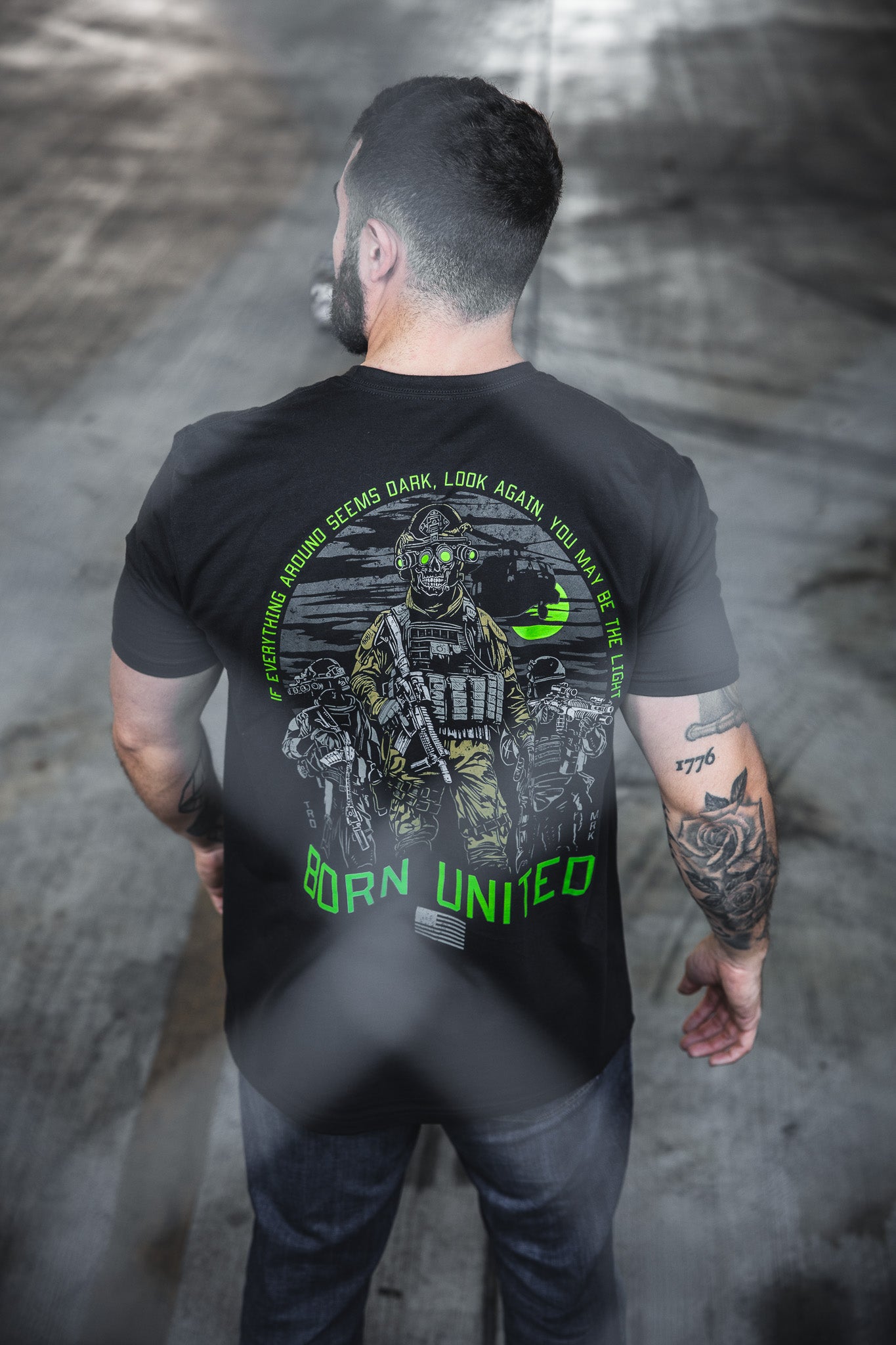 A tattooed man, seen from behind, wears the Born United Private Label “Light in the Darkness” black 60/40 tee with soldier graphic, green accents, and the quote: If everything around seems dark, look again—you may be the light.
