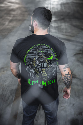 A tattooed man, seen from behind, wears the Born United Private Label “Light in the Darkness” black 60/40 tee with soldier graphic, green accents, and the quote: If everything around seems dark, look again—you may be the light.