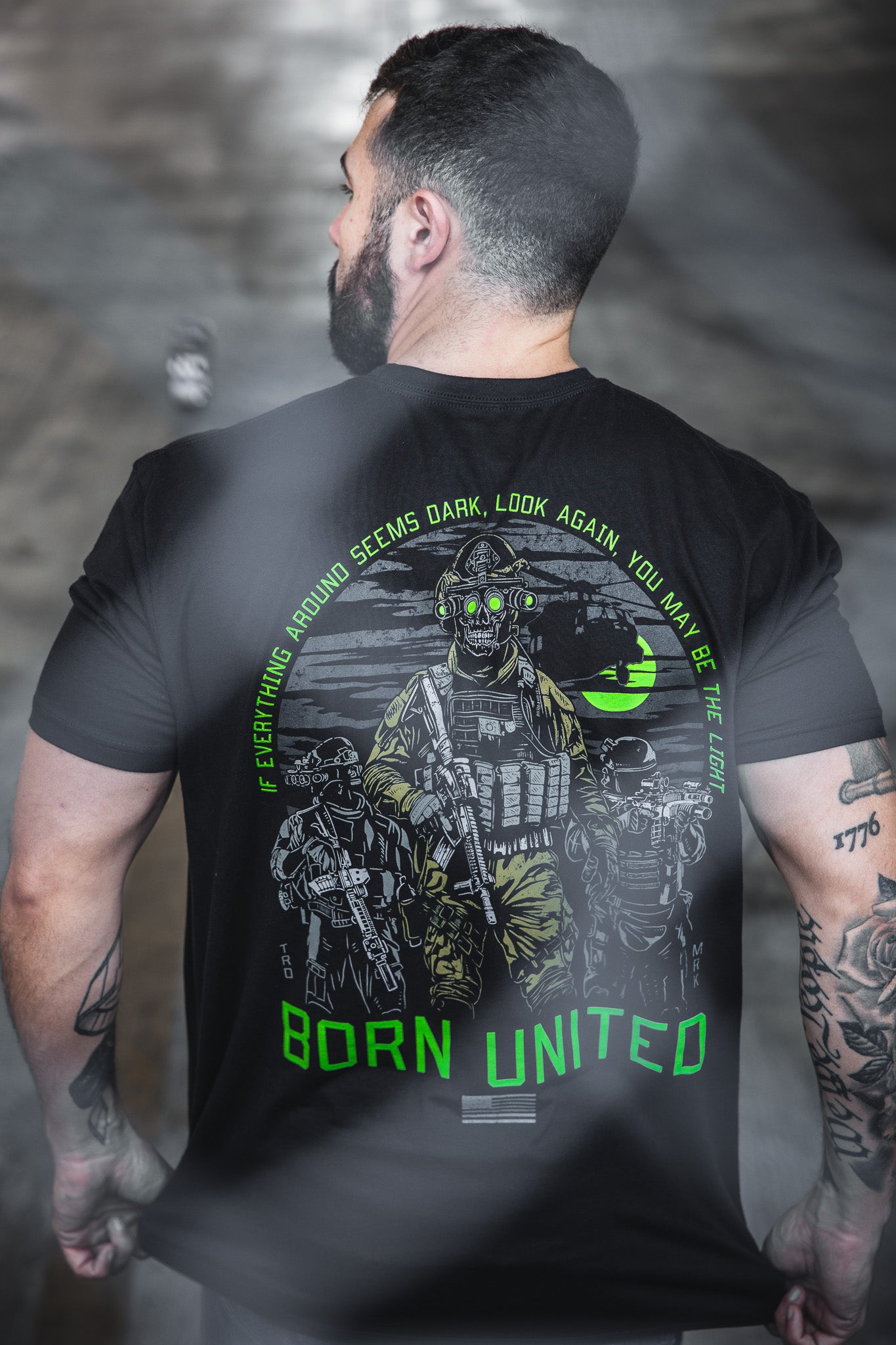 A tattooed man faces away, wearing the Light in the Darkness black 60/40 tee by Born United Private Label, featuring three armored soldiers, neon green accents, “Born United,” and the motto: "If everything around seems dark, look again, you may be the light.
