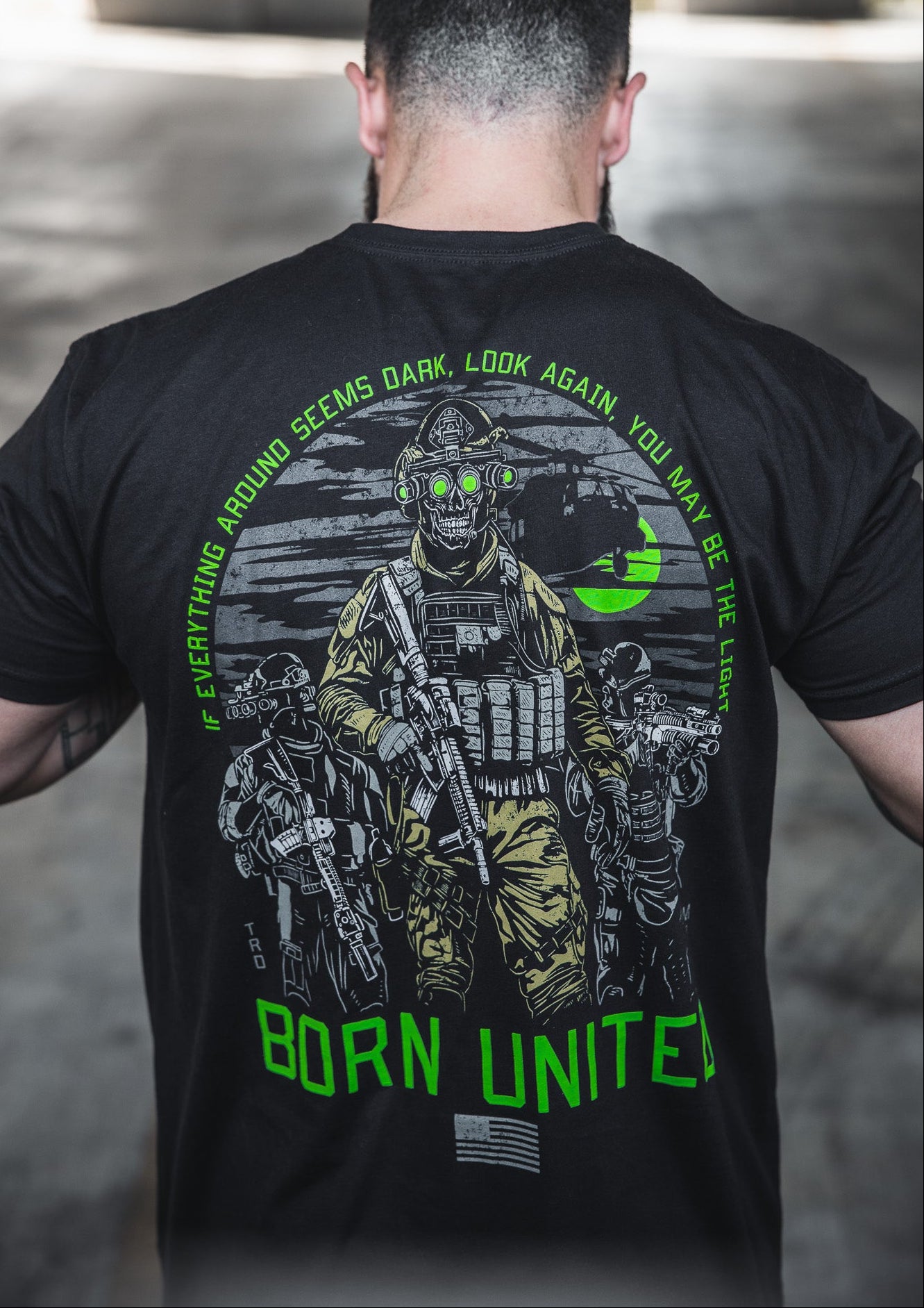 A bearded man faces away, wearing Born United Private Label's "Light in the Darkness" black 60/40 blend shirt with armed soldiers in night vision. The shirt reads: "Everything around seems dark, look again, you may be the light" and Born United.