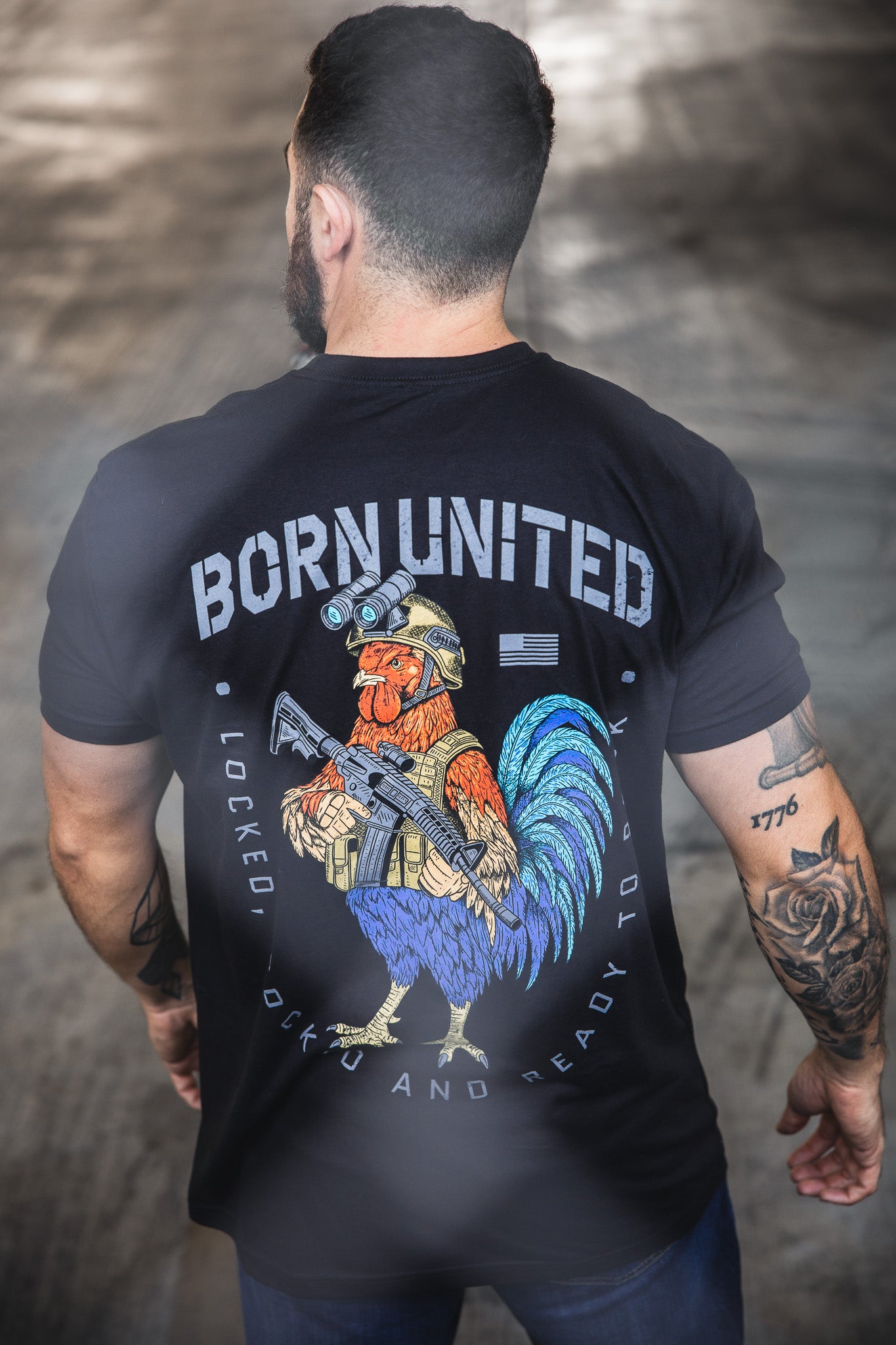 A tattooed man wears a black Battle Bird T-shirt from Born United Private Label, showing a colorful rooster in military gear with a rifle and the slogans “BORN UNITED” and “Locked, Cocked and Ready to Rock,” standing with his back to the camera.