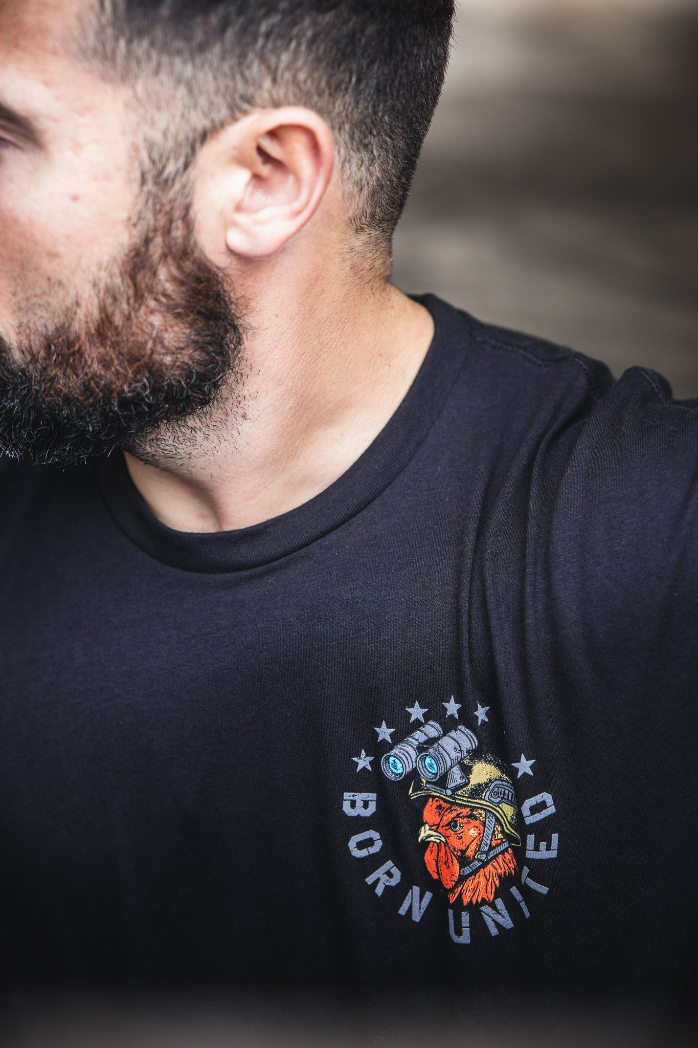 A man with a beard wears a dark 60/40 blend Born United Private Label Battle Bird t-shirt, featuring a dog in a helmet with night vision goggles, stars, and “BORN UNITED.” The image highlights the shirt’s design.