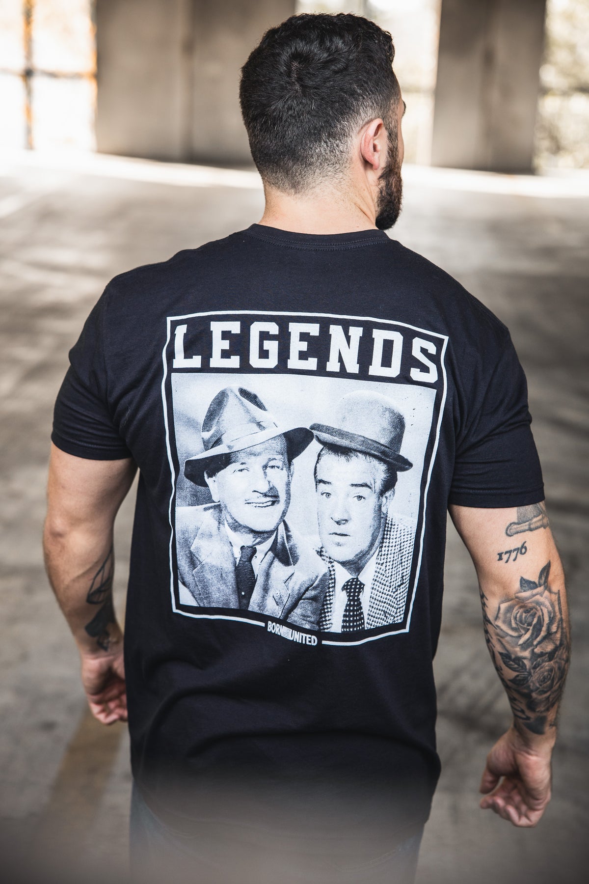 A tattooed man stands with his back to the camera, wearing a Born United Private Label "Legends - Bud and Lou" black T-shirt featuring vintage photos of two men in suits and hats, with LEGENDS printed above them.