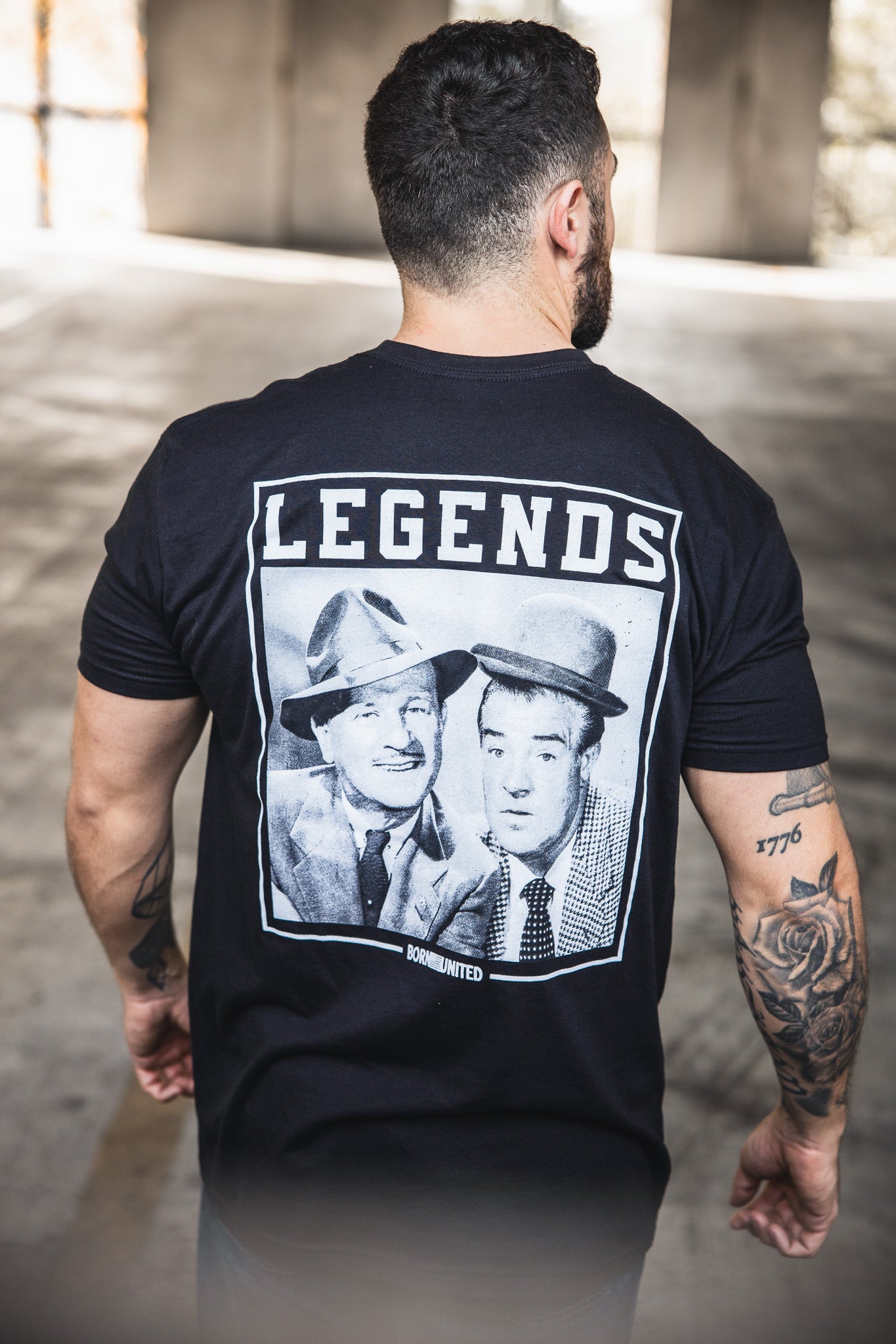 A tattooed man stands with his back to the camera, wearing a Born United Private Label "Legends - Bud and Lou" black T-shirt featuring vintage photos of two men in suits and hats, with LEGENDS printed above them.