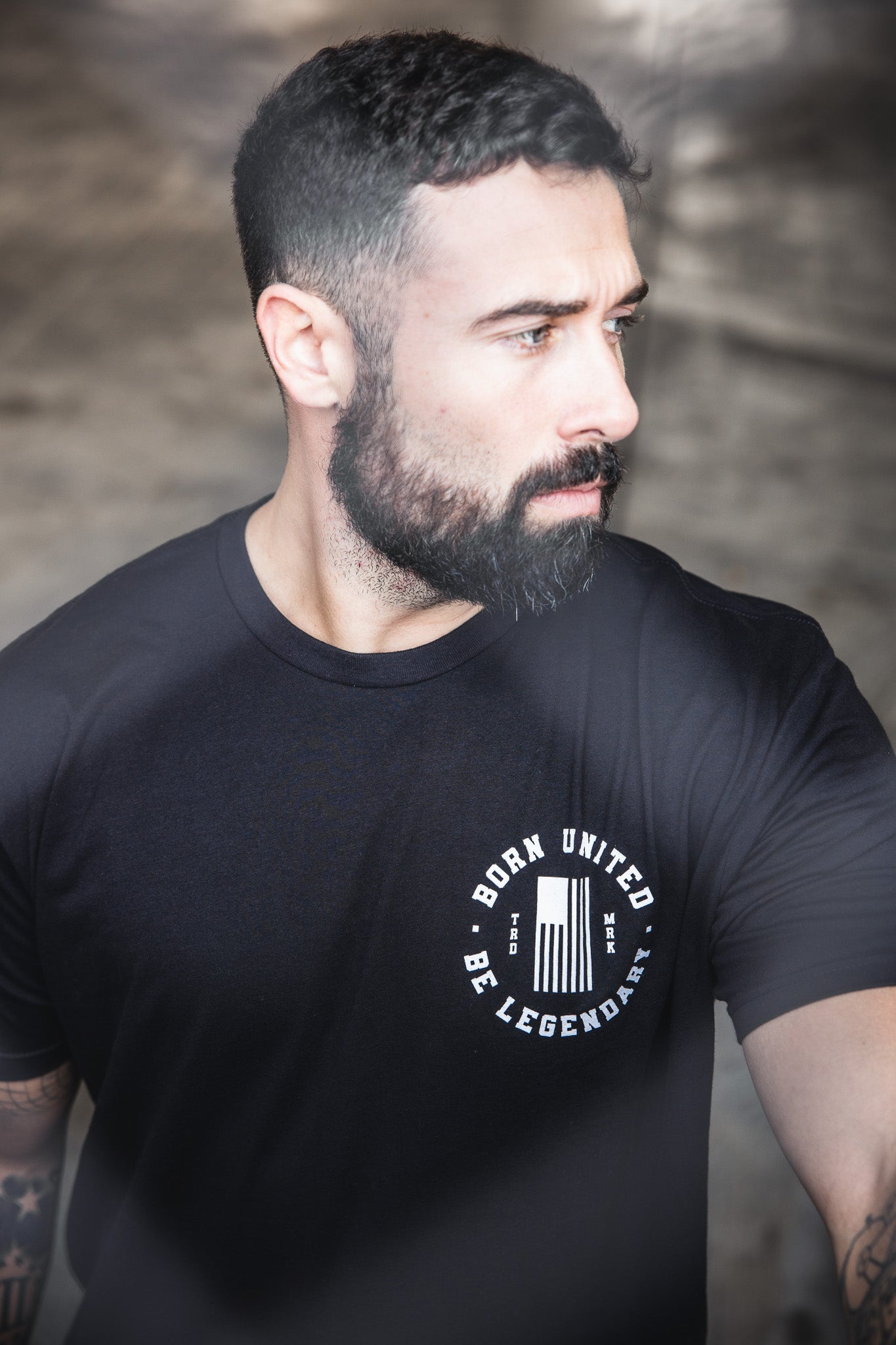A bearded man with short dark hair wears a black "Legends - Bud and Lou" T-shirt by Born United Private Label and looks thoughtfully to the side outdoors.
