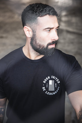 A bearded man with short dark hair wears a black "Legends - Bud and Lou" T-shirt by Born United Private Label and looks thoughtfully to the side outdoors.