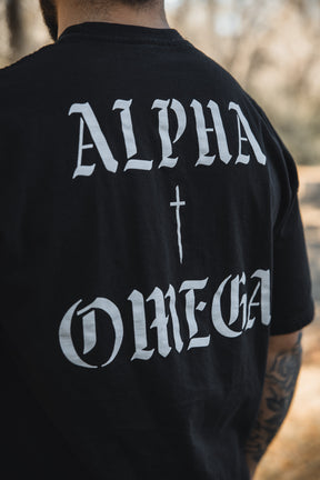 A person wears the 13th Alpha & Omega Premium Oversized Tee in black—an oversized streetwear look with bold white gothic “ALPHA” and “OMEGA” lettering flanking a cross. Back view highlights only part of one tattooed arm—Christian-inspired style.