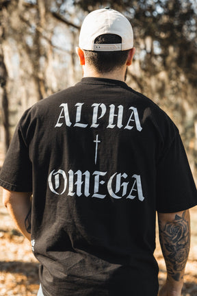 Wearing the 13th Alpha & Omega Premium Oversized Tee in black, featuring ALPHA and OMEGA divided by a cross on the back, a person stands outdoors among blurred trees, paired with a white cap.